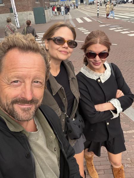 This vibrant selfie captures three friends enjoying a sunny afternoon on Raadhuisstraat in Centrum, Amsterdam, with historic brick buildings and a bustling street scene in the background